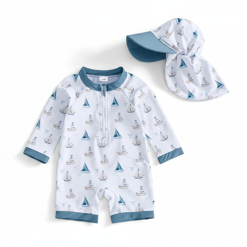 Baby and toddler boy one-piece long sleeve rash guard swimsuit featuring a watercolor sailboat and whale tail print on ivory fabric, shown on a white background with a matching wide-brim sun hat.