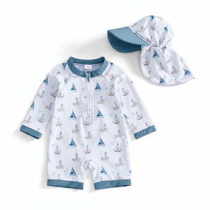 Baby and toddler boy one-piece long sleeve rash guard swimsuit featuring a watercolor sailboat and whale tail print on ivory fabric, shown on a white background with a matching wide-brim sun hat.