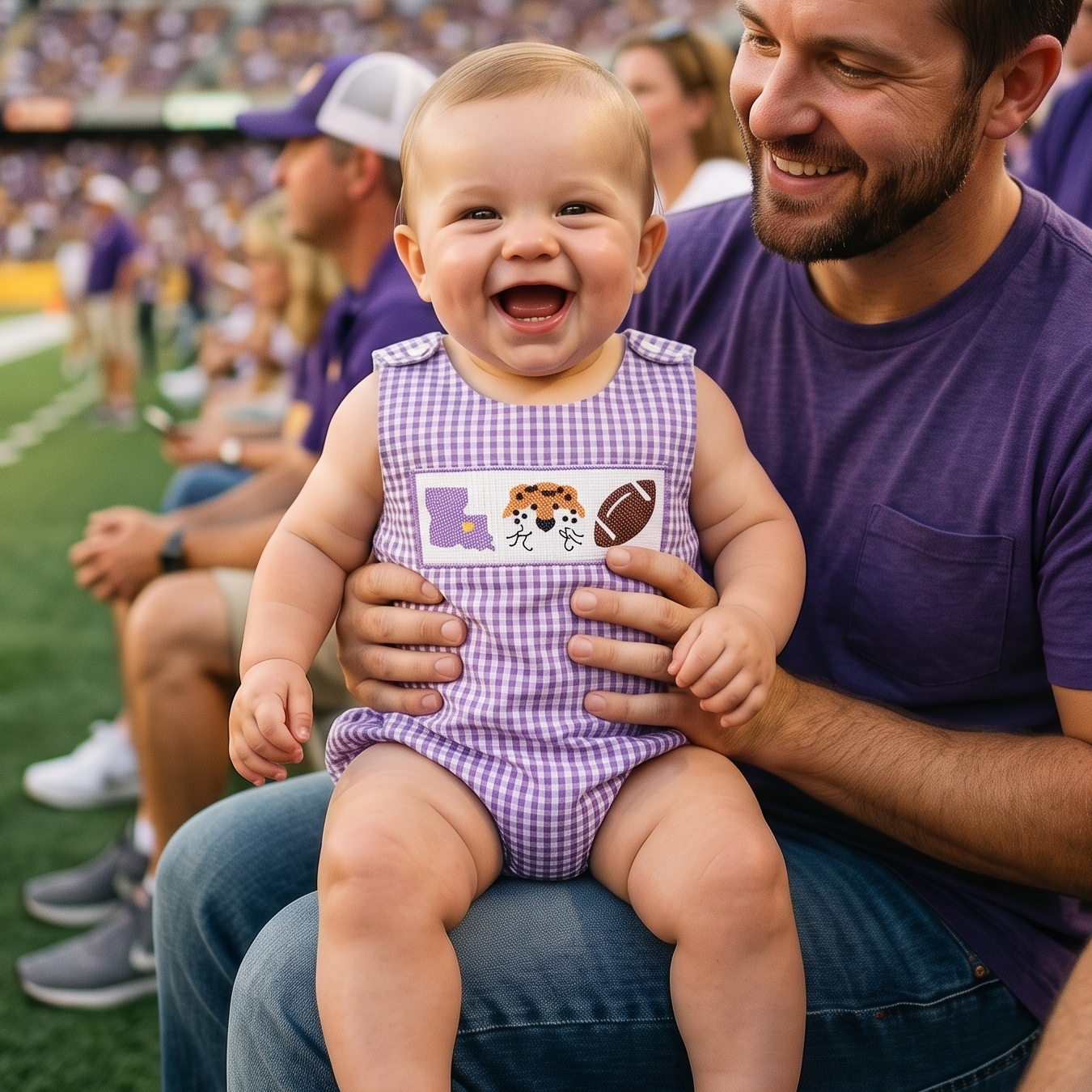 A smiling baby boy sitting on his dad's lap in stadium bleachers, wearing a purple gingham smocked bubble romper featuring an embroidered purple state outline, tiger face, and football.