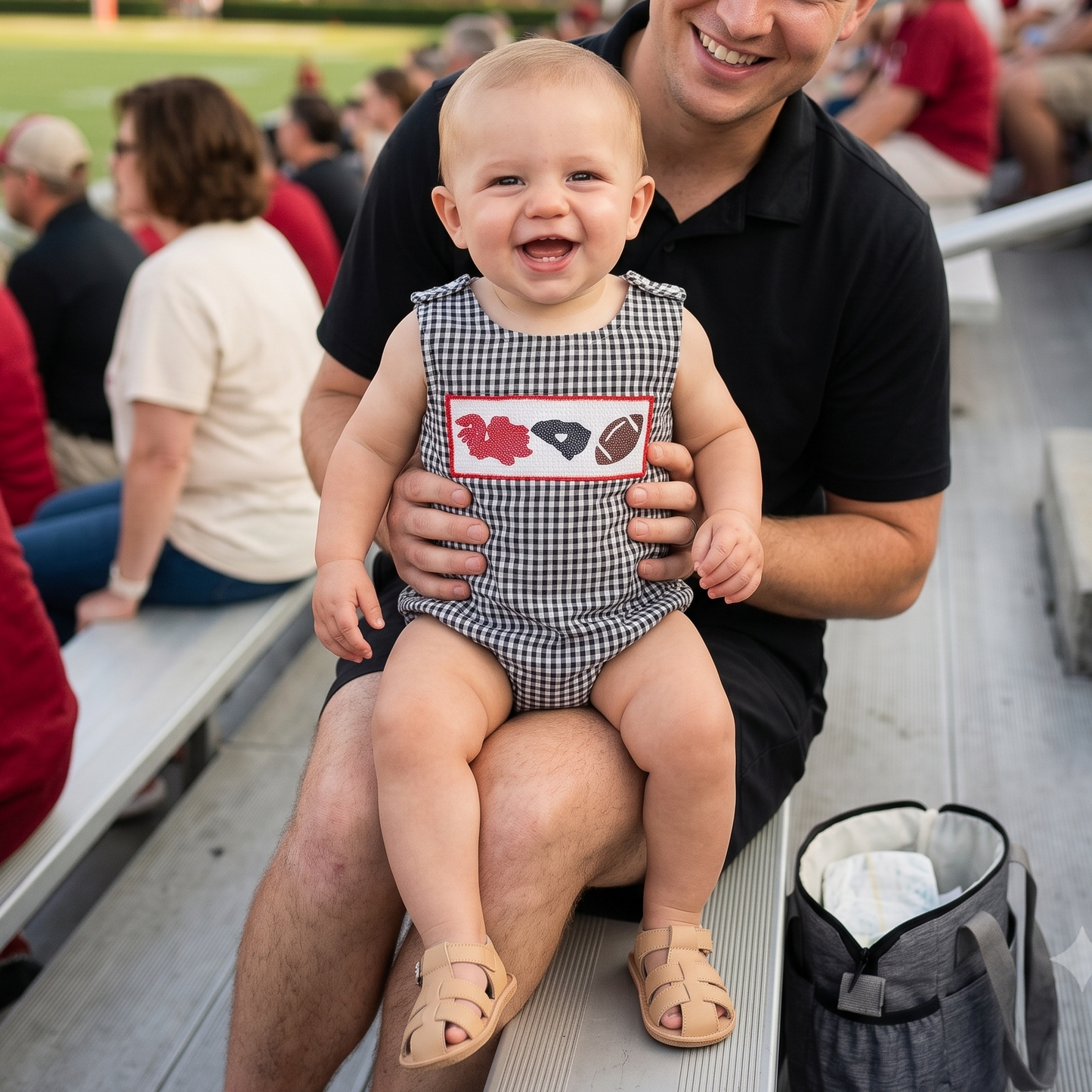 A smiling baby boy sitting on his dad's lap in stadium bleachers, wearing a black gingham smocked bubble romper featuring embroidered game day details, a football, and a garnet state outline.
