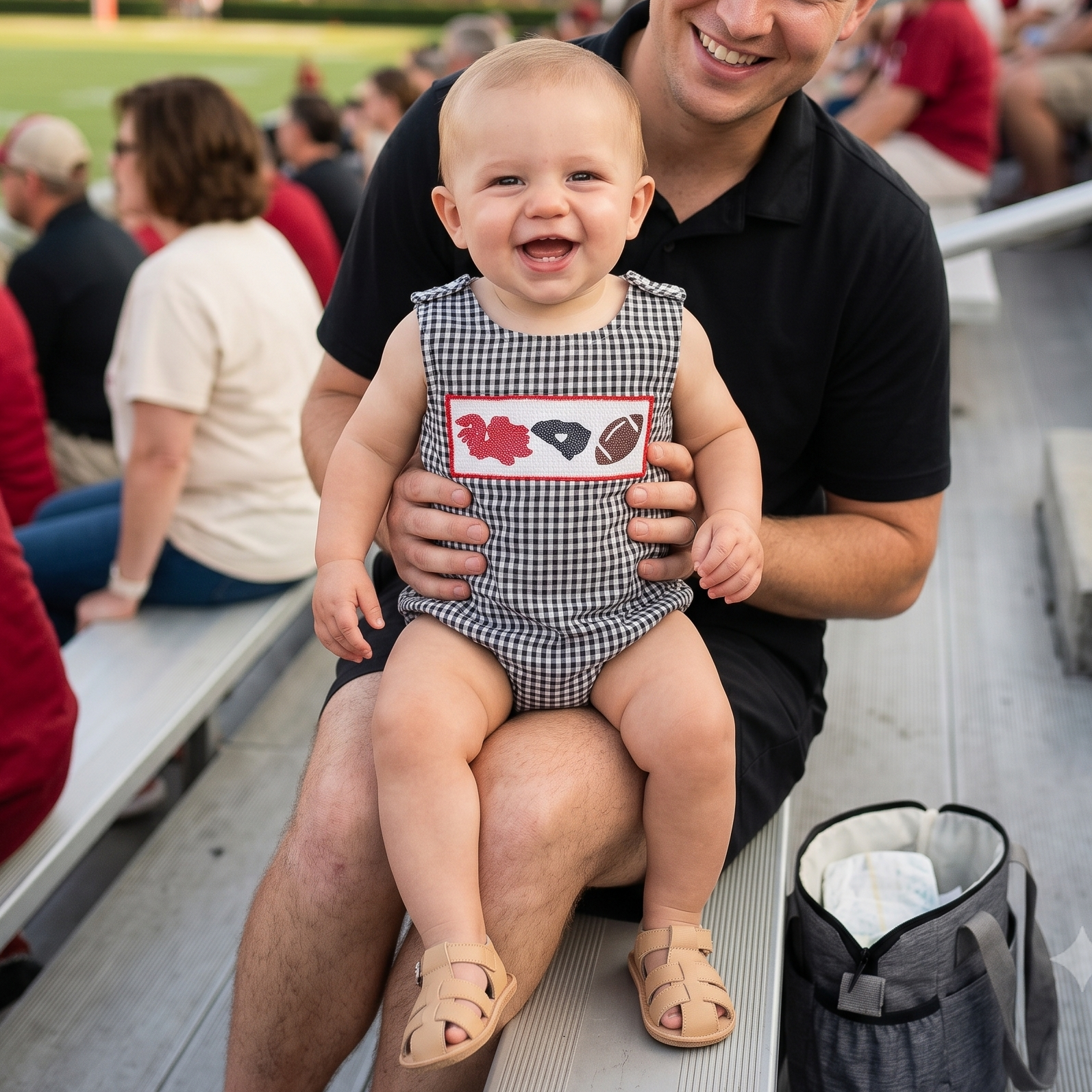 A smiling baby boy sitting on his dad's lap in stadium bleachers, wearing a black gingham smocked bubble romper featuring embroidered game day details, a football, and a garnet state outline.