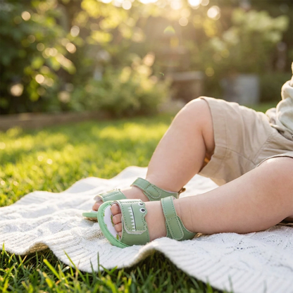 Baby sitting on a white blanket outdoors wearing green soft sole crocodile first walker sandals.