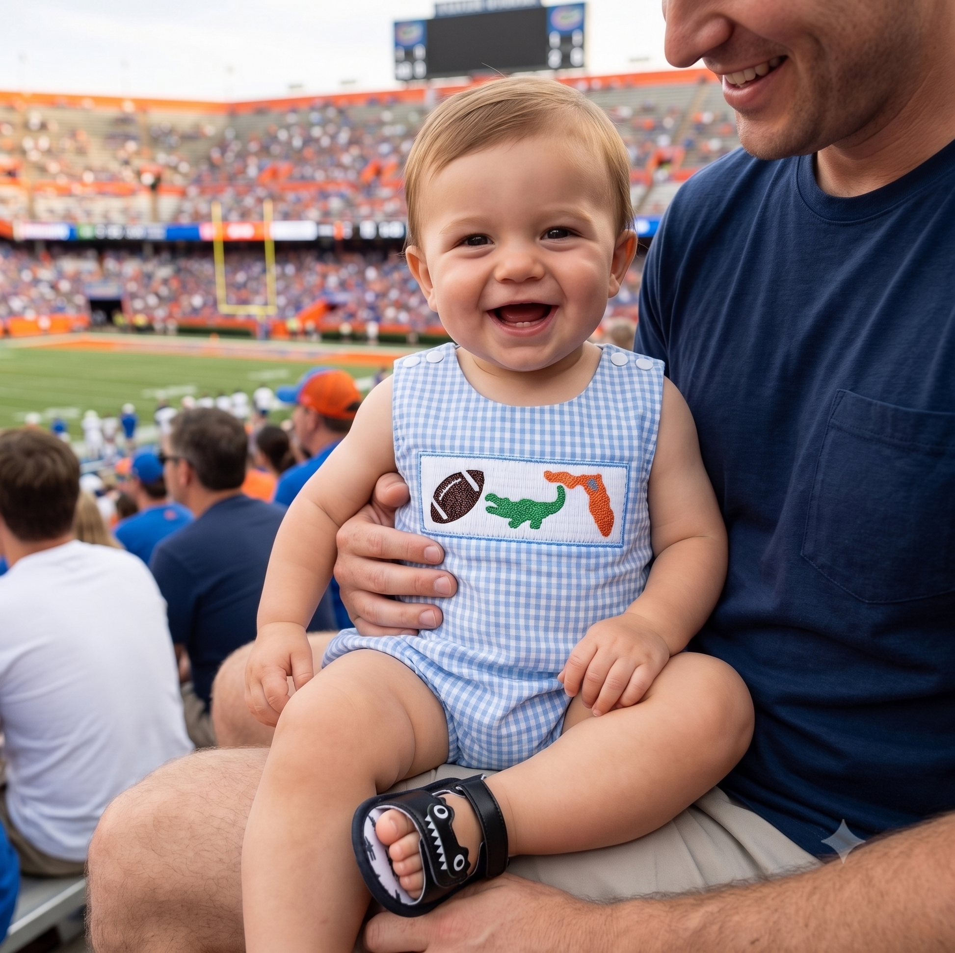 A smiling baby boy sitting on his dad's lap in stadium bleachers during a game, wearing a light blue gingham smocked bubble romper featuring embroidered football, green alligator, and orange state outline details.