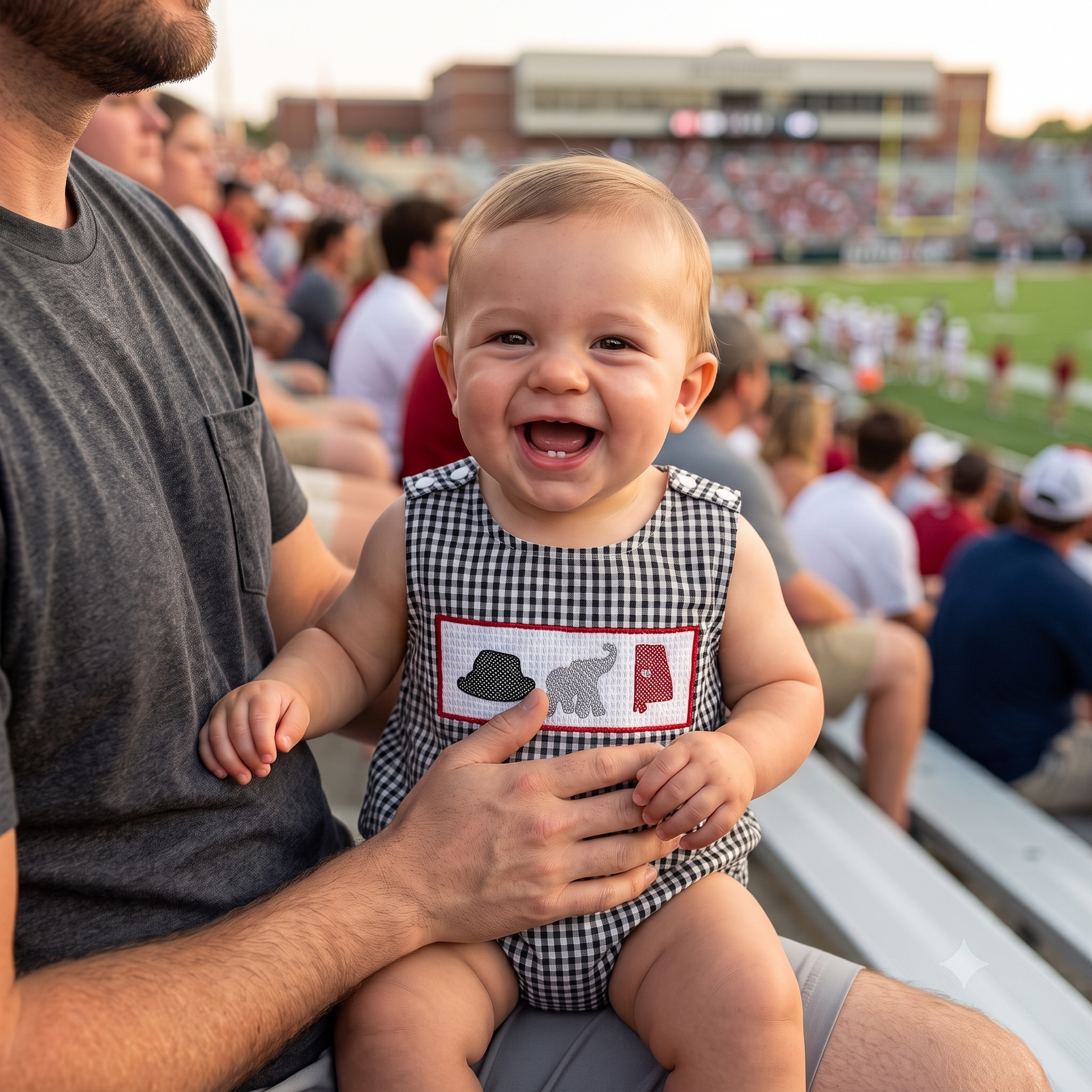 A smiling baby boy sitting in stadium bleachers with dad during a college football game, wearing a black gingham smocked bubble romper with a crimson state outline, houndstooth hat, and grey elephant embroidery.