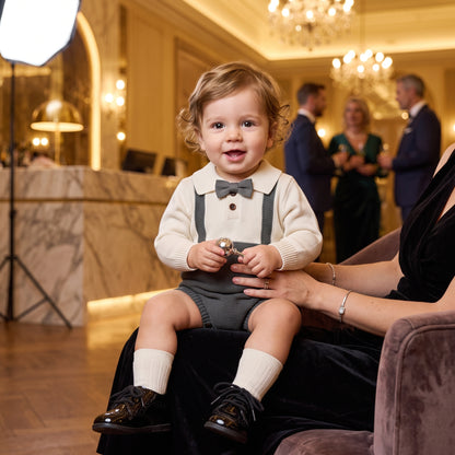 A happy baby boy wearing a formal gray and cream knit suspender romper with a bow tie at a special event or wedding.