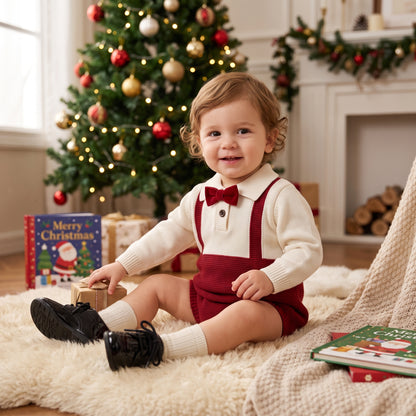 A happy baby boy sitting on a fluffy rug in front of a Christmas tree, wearing a red and cream knit suspender romper with a bow tie.