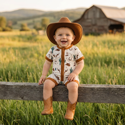 A smiling baby boy wearing a cream western romper, brown cowboy hat, and cowboy boots, sitting on a rustic wooden fence in a green field.