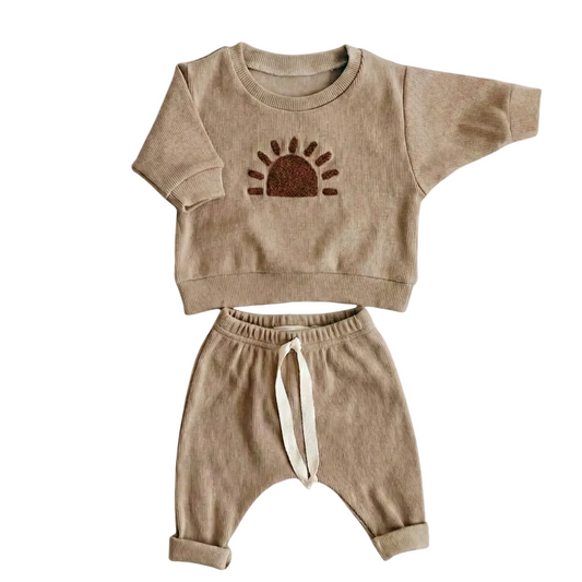 A flat lay photograph of a two-piece beige ribbed knit toddler outfit. The set includes a long-sleeved pullover sweatshirt with a brown embroidered sun design on the chest and matching drawstring pants with cuffed ankles, all laid against a plain light beige background.