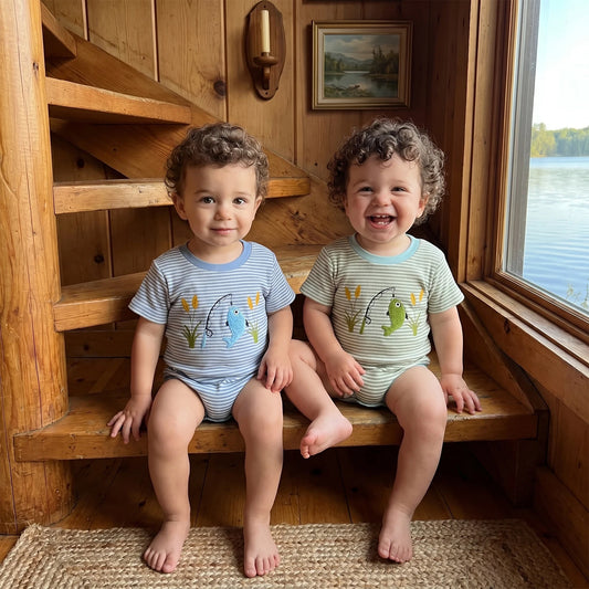 Two smiling baby boys sitting on rustic wooden stairs in a lake house setting, wearing the blue and green striped short-sleeve fishing rompers.