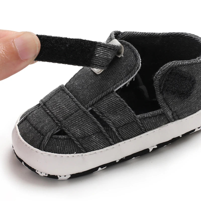 Close-up of a hand opening the adjustable velcro strap on a dark gray black baby canvas summer sandal.