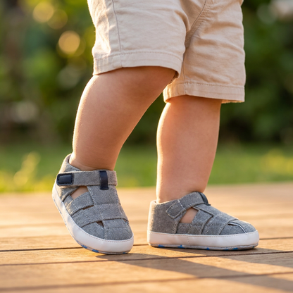 A toddler standing outdoors on a wooden deck wearing light gray canvas summer sandals.