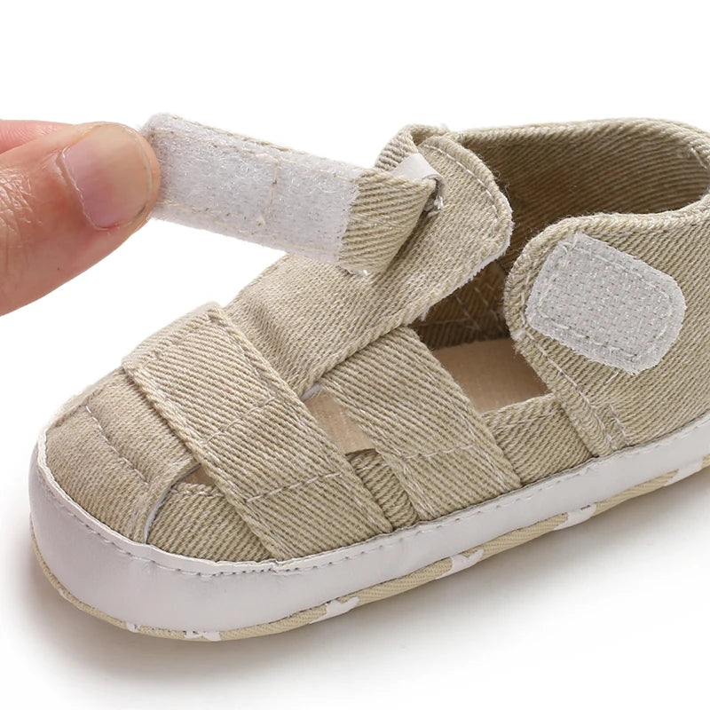 Close-up of a hand opening the adjustable velcro strap on a tan beige baby canvas summer sandal.