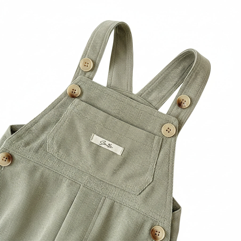 Close-up of button shoulder straps, front pocket, and cursive Smile chest tag on sage green corduroy overalls.