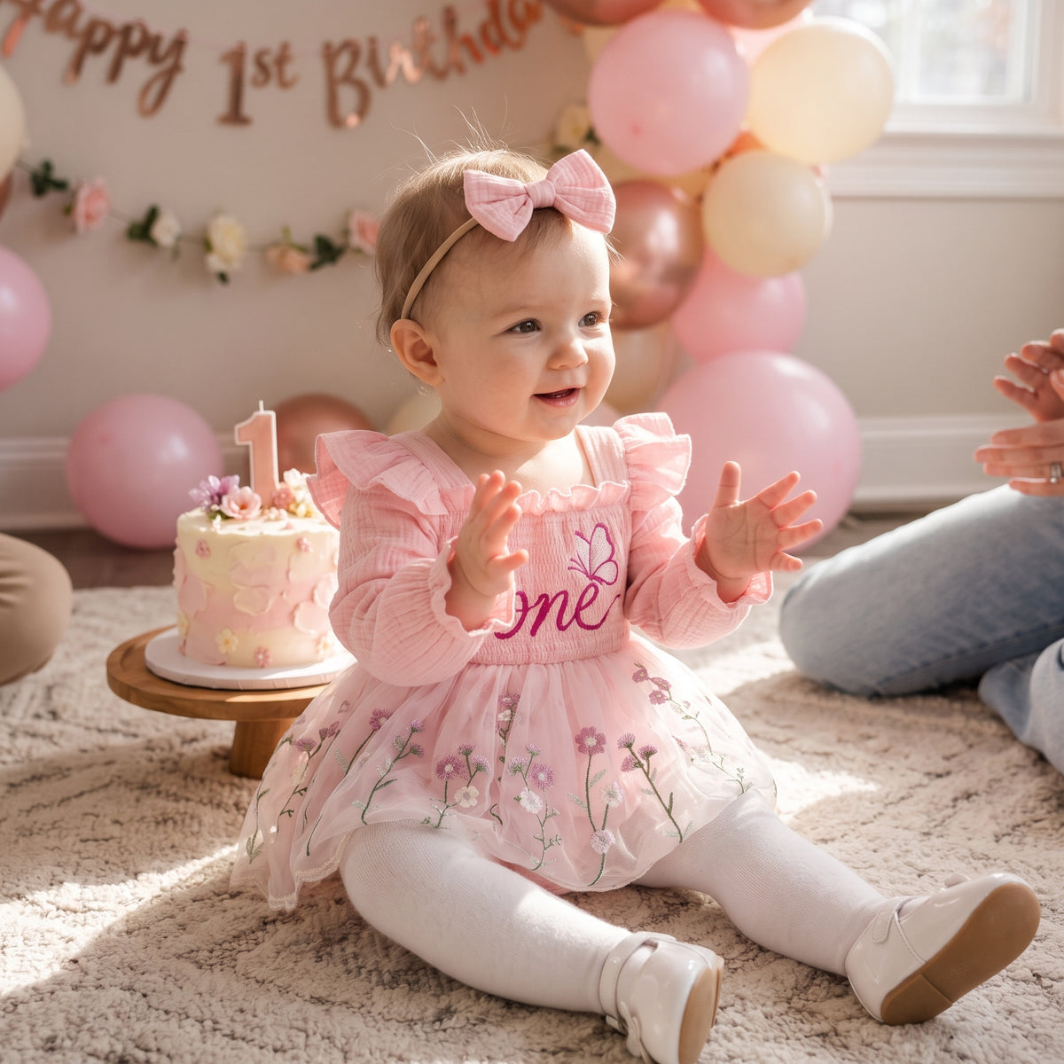 A baby girl sitting on a beige rug at a first birthday party, wearing a pink long-sleeve dress with 'one' embroidered on the chest, white tights, and a pink bow headband. A small pink and white cake with a candle is on a stand nearby, with pink and cream balloons and a 'Happy 1st Birthday' banner in the background.