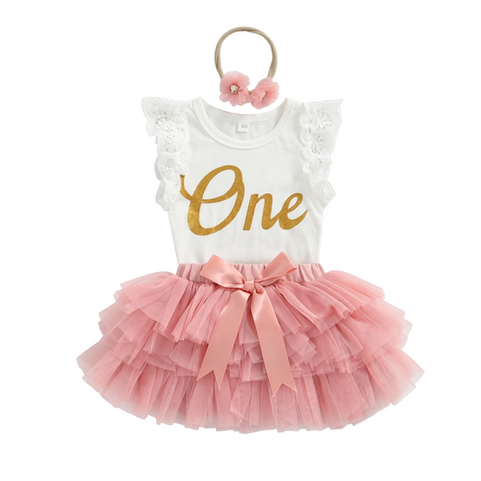 Flat lay of baby girl 1st birthday 3-piece outfit featuring a white lace romper with gold glitter "One" print, a pink multi-layered tulle tutu skirt with a satin bow, and a matching floral headband on a white background.