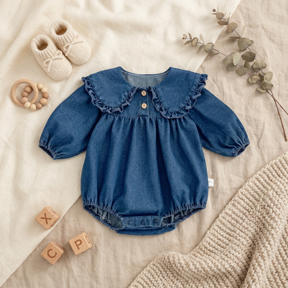 Styled flat lay of a baby girl blue denim long-sleeve romper with a ruffle Peter Pan collar, surrounded by cream knit baby booties, wooden blocks, a wooden teether, and a sprig of eucalyptus on a textured neutral background.