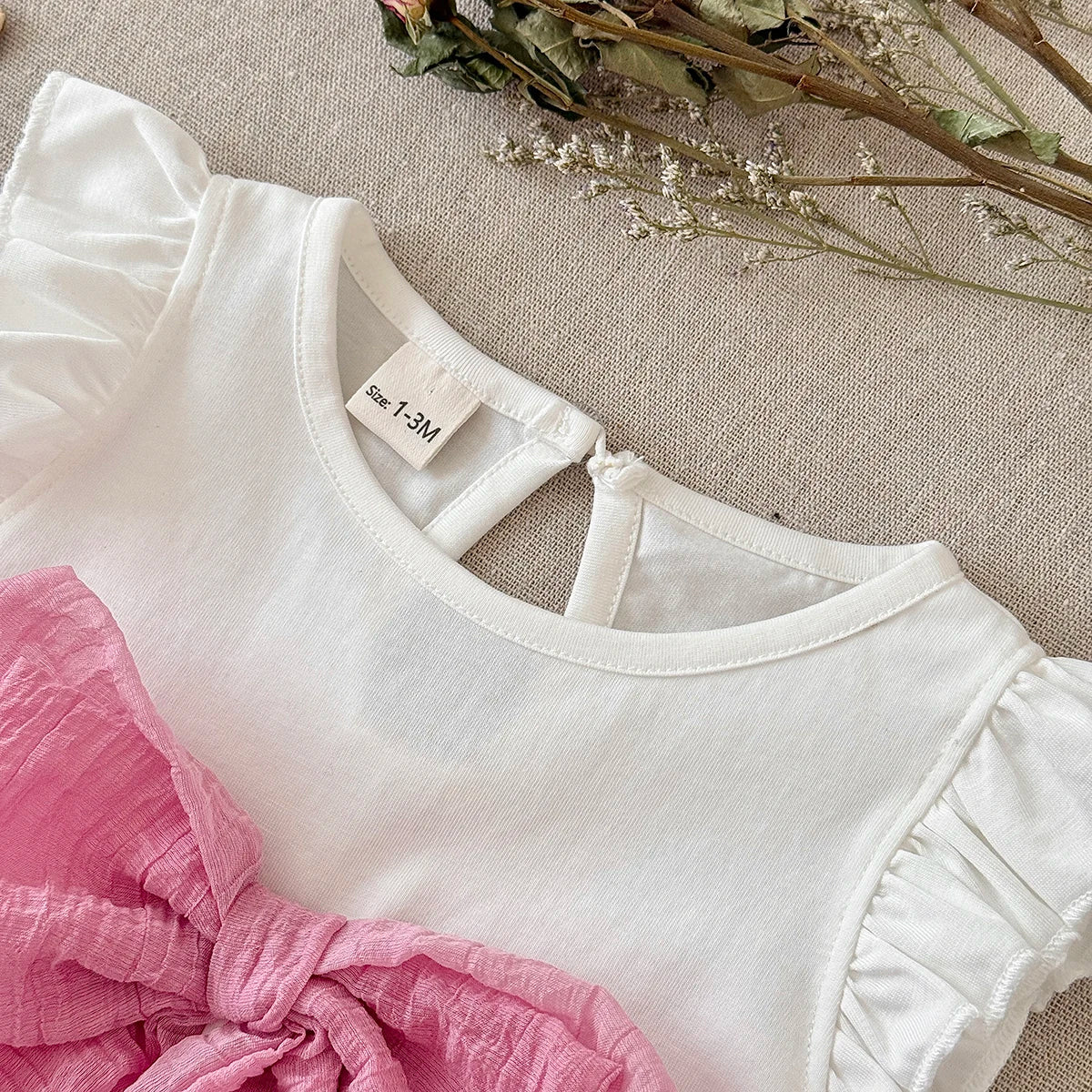 Close-up view of the white bodysuit's crew neckline, rear keyhole button closure, ruffle shoulder details, and the textured pink front bow.
