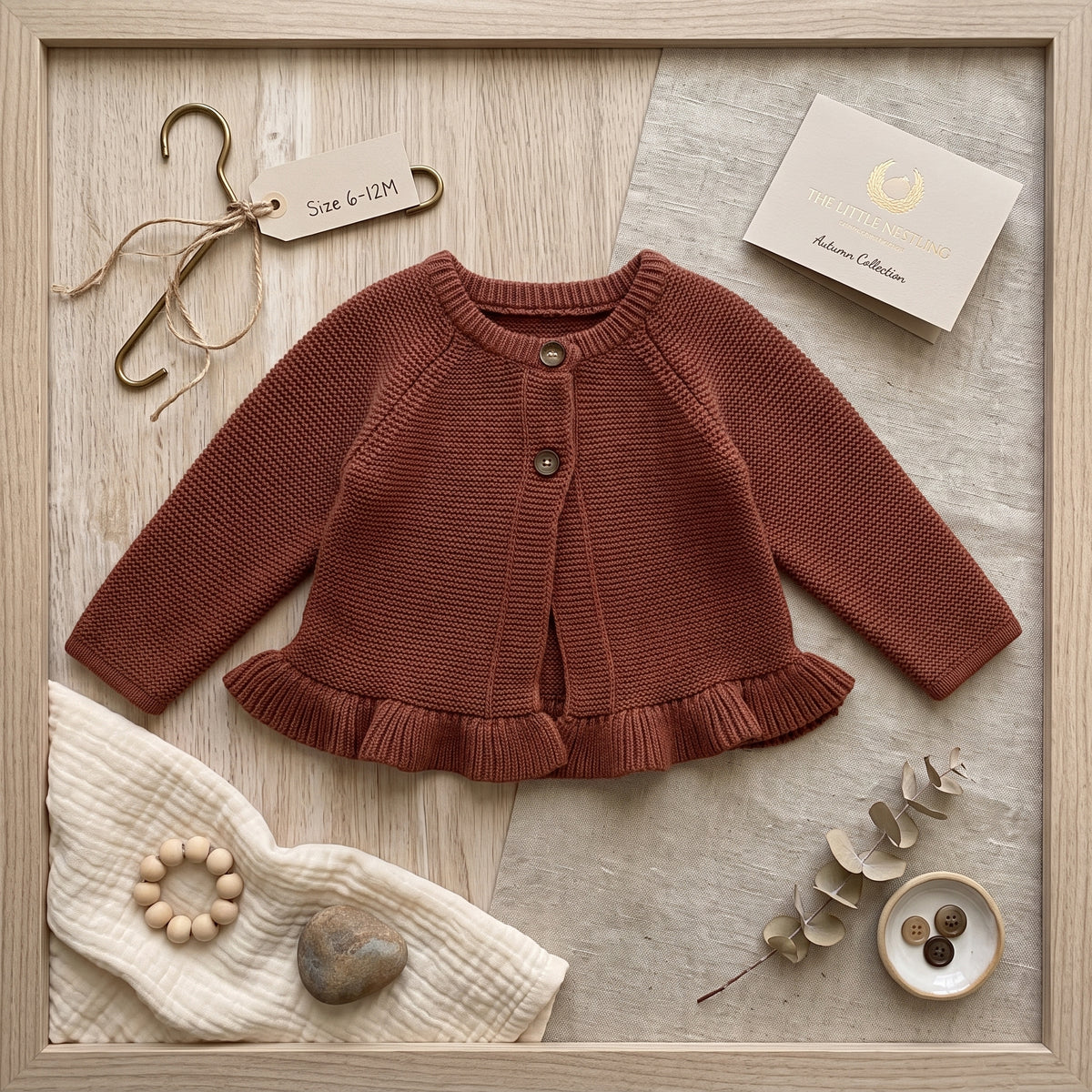 Flat lay of a baby girl's brown knit sweater with a ruffled collar and hem, styled aesthetically next to a wooden hanger, a teething ring, and decorative dried foliage.