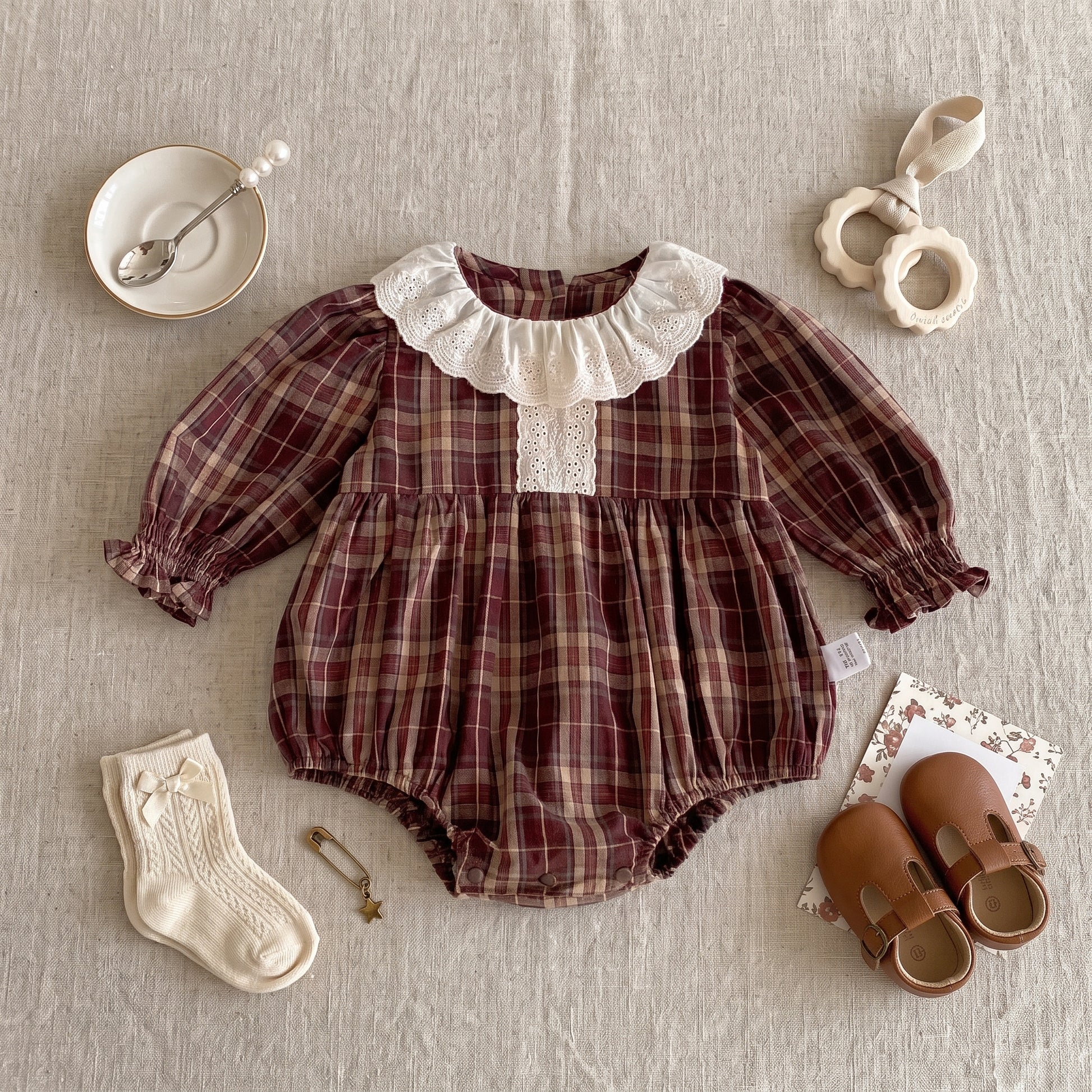 Flat lay of a long sleeve brown plaid baby girl romper styled with cream socks, leather shoes, and accessories.