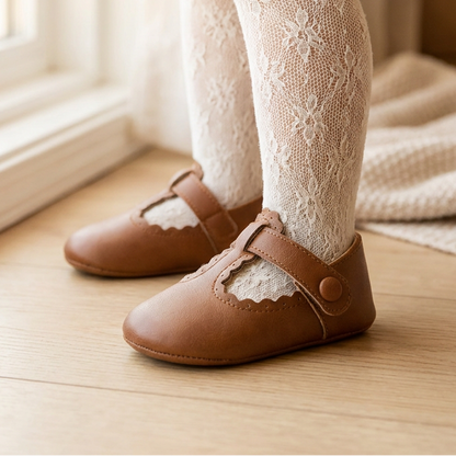 Toddler wearing brown PU leather T-strap Mary Jane shoes and lace tights, standing on a wooden floor.