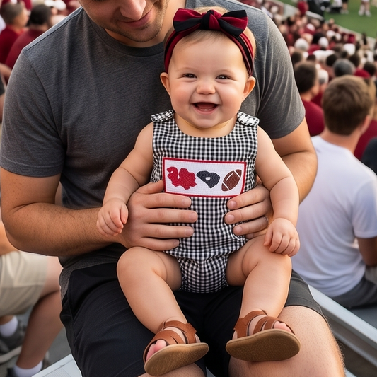 A smiling baby girl wearing a garnet bow headband sitting on her dad's lap in stadium bleachers, dressed in a black gingham smocked bubble romper featuring embroidered game day details and a garnet state outline.