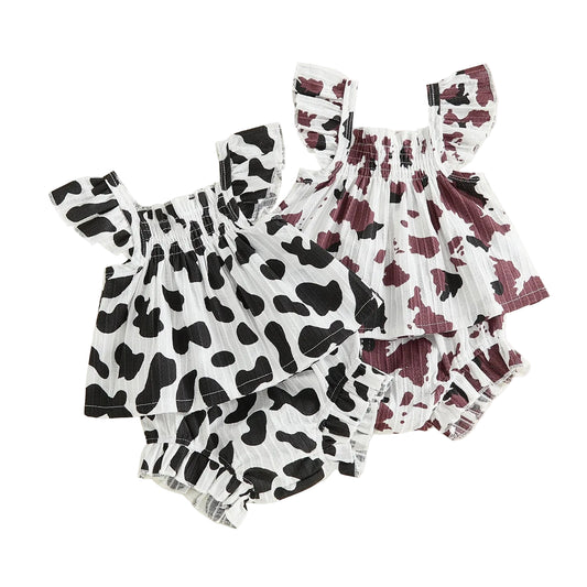 tudio shot displaying two baby girl summer outfits featuring black and white, and brown and white cow prints with smocked ruffled tops and matching shorts.