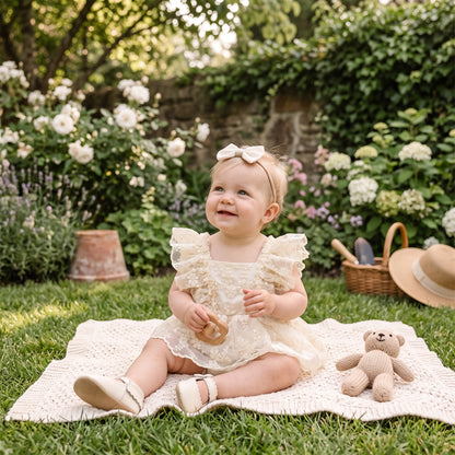 Baby girl wearing cream floral lace romper dress outdoors on garden picnic blanket