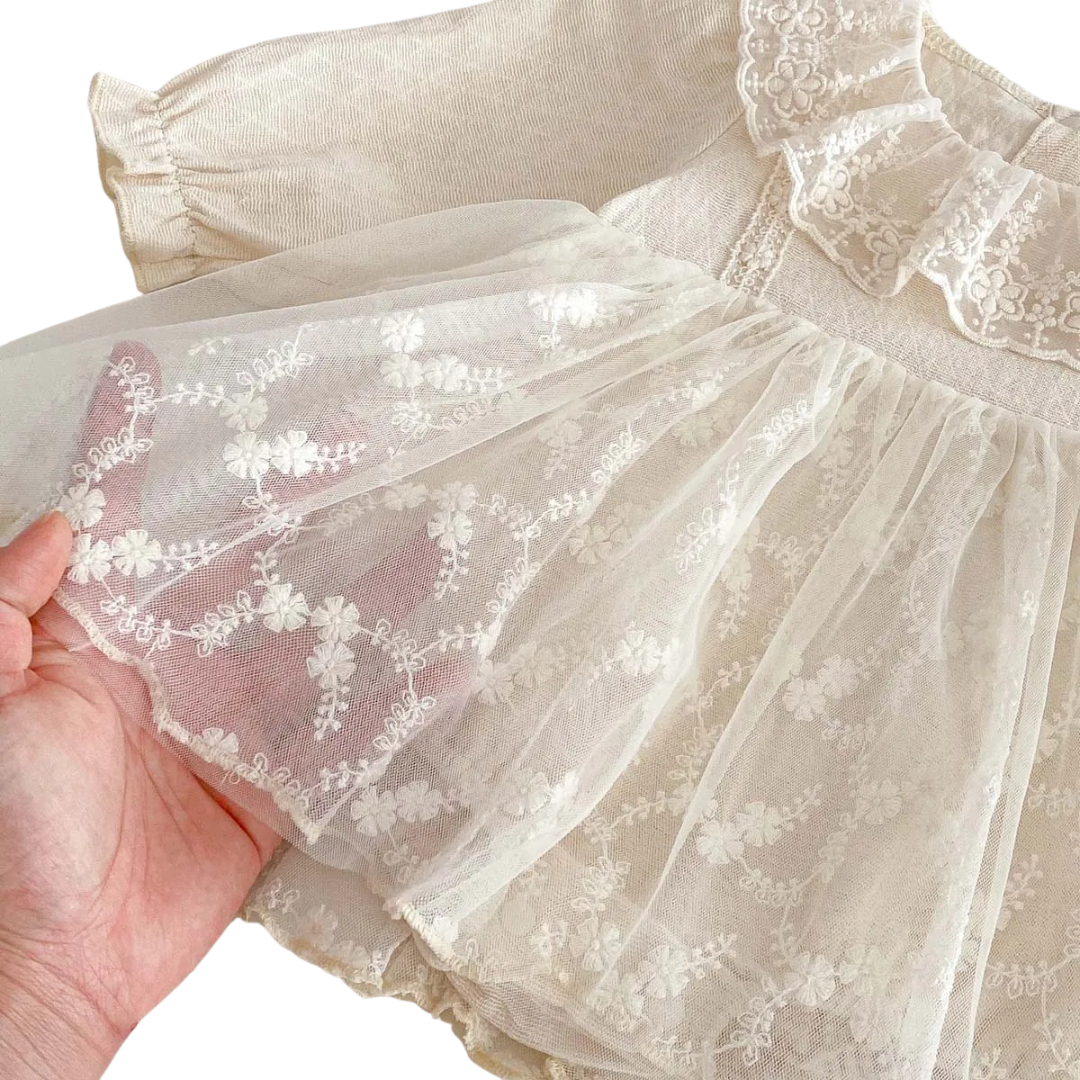 A hand gently lifting the sheer tulle skirt overlay of the baby girl dress to show the intricate floral embroidery.