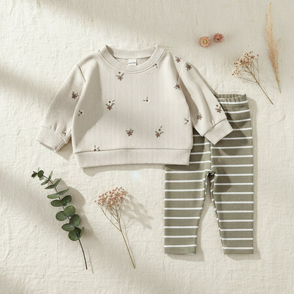 Styled flat lay of a baby girl's floral ribbed sweatshirt and striped pants set on a textured cream background with dried botanical props.