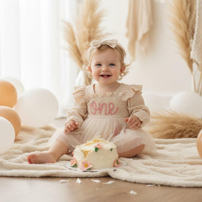 Baby girl wearing cream smocked first birthday outfit sitting next to a birthday cake for a photo shoot