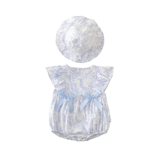 Flat lay of a baby girl's light blue and white floral cotton romper with ruffled sleeves and two blue satin bows, paired with a matching wide-brim sun hat on a neutral textured background.