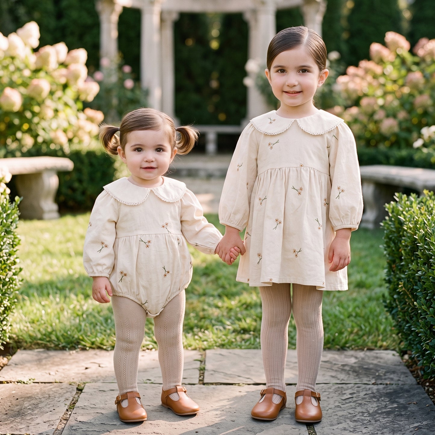 Baby girl wearing cream floral embroidered romper holding hands with older sister wearing matching dress outdoors