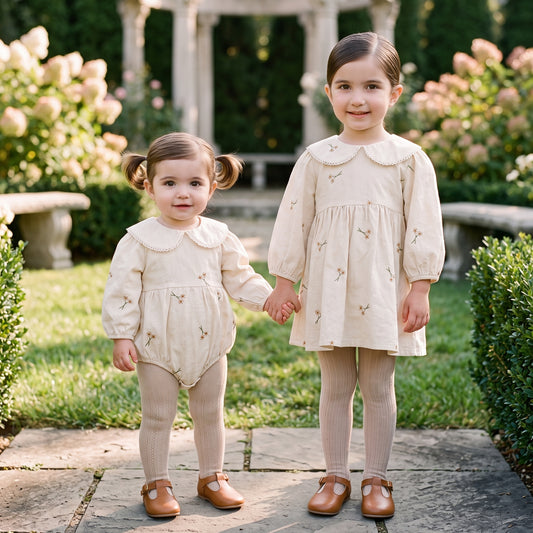 Baby girl wearing cream floral embroidered romper holding hands with older sister wearing matching dress outdoors