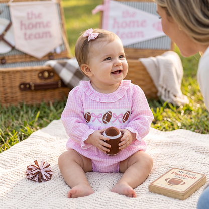 Smiling baby girl wearing a pink smocked football romper sitting on a blanket outdoors for a fall tailgate.