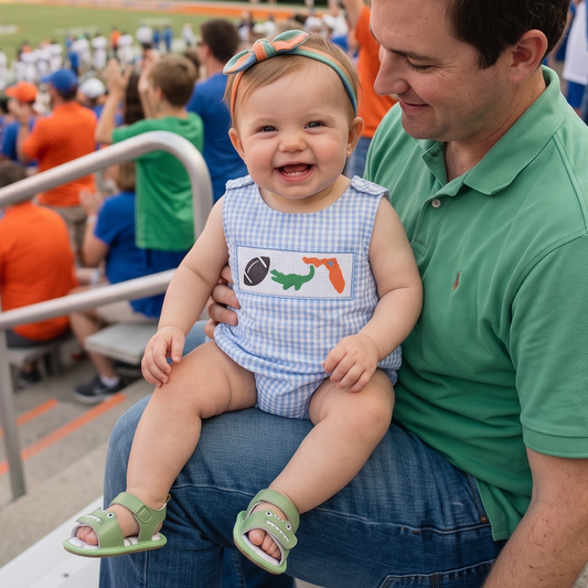 A smiling baby girl sitting on her dad's lap in stadium bleachers, wearing a light blue gingham smocked bubble romper featuring embroidered football, green alligator, and orange state outline details.