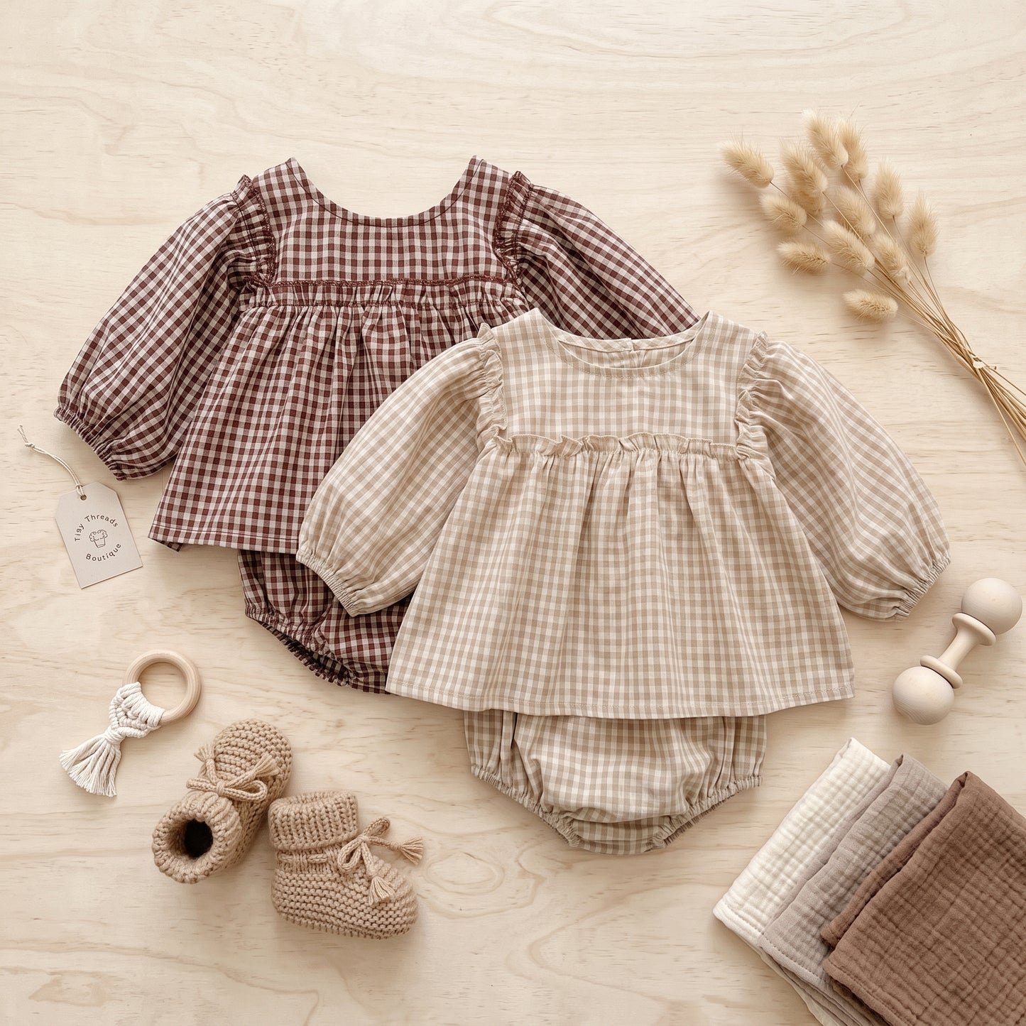 Flat lay view of beige and brown baby girl smocked gingham 2-piece outfit sets with booties and wooden rattle.