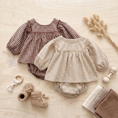 Flat lay view of beige and brown baby girl smocked gingham 2-piece outfit sets with booties and wooden rattle.