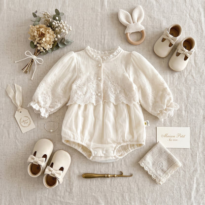 Flat lay of ivory vintage-inspired baby girl lace romper styled with baby shoes, wooden teether, and floral accents.