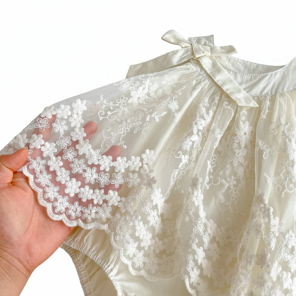 Close-up view of the detailed floral lace embroidery and the elegant satin shoulder bow on the Pearl Ivory baby girl romper dress.