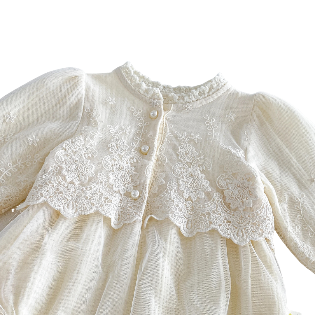 Close-up of scalloped embroidered lace trim and pearl button details on an ivory long sleeve baby bodysuit.