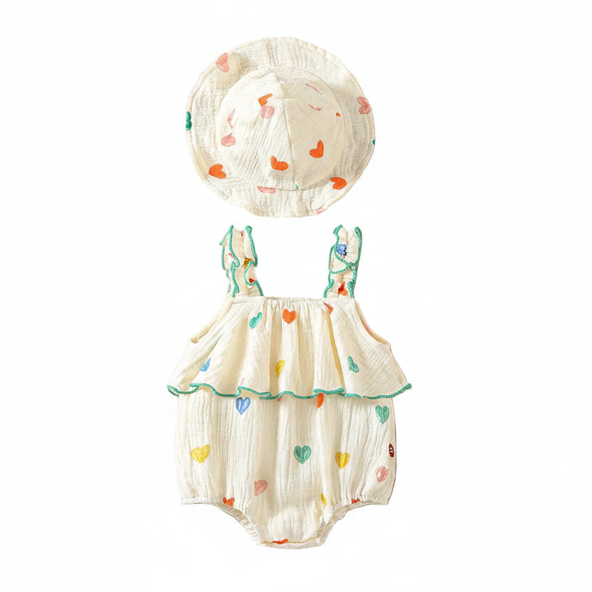 Flat lay of a white muslin cotton baby girl romper with colorful heart prints, ruffle straps, and a matching wide-brim sun hat on a clean white background.