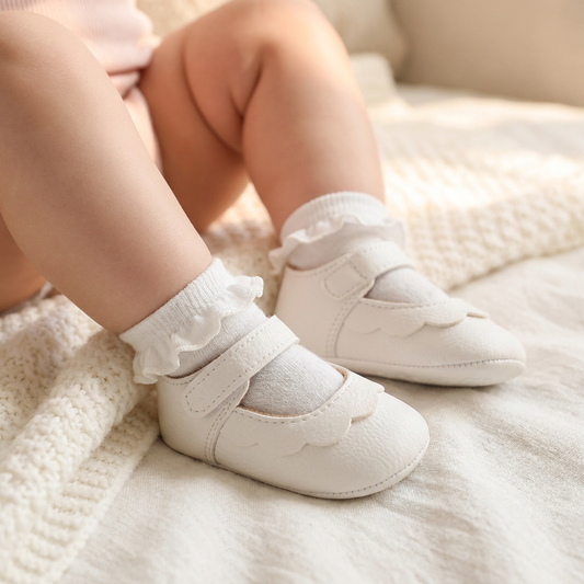 Close-up lifestyle shot of an infant girl wearing white scalloped Mary Jane shoes and ruffled socks, sitting on a soft cream knit blanket in a sunlit nursery.