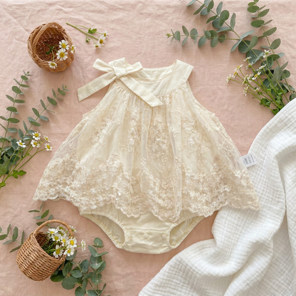 Styled spring lifestyle flat lay of the Pearl Ivory baby girl lace romper dress on a soft pink linen background surrounded by fresh daisies, eucalyptus branches, and small wicker baskets.