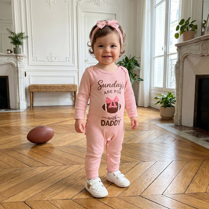 Happy baby girl standing on a hardwood floor indoors next to a football, wearing the pink ribbed "Sundays With Daddy" long sleeve romper.