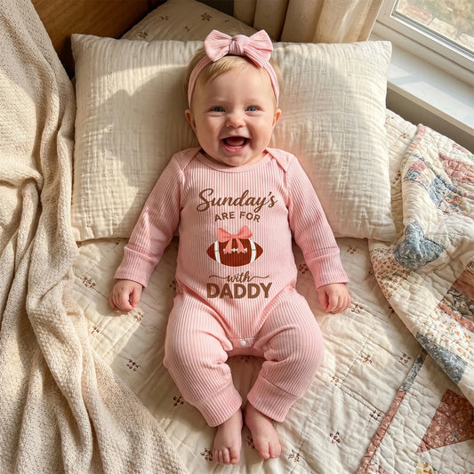 Smiling baby girl lying on a cozy bed wearing a pink ribbed "Sundays With Daddy" football romper and matching headband.