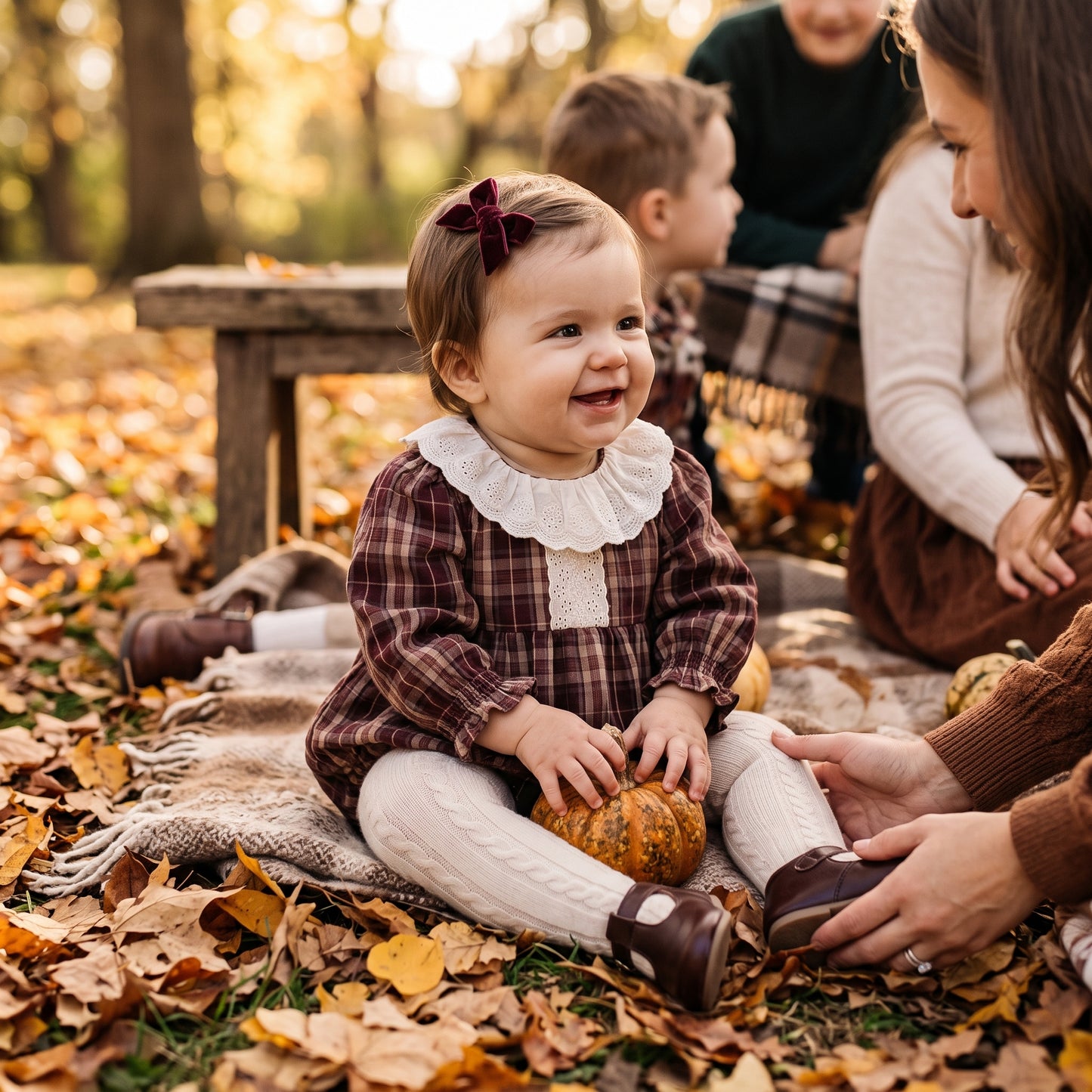 Baby girl wearing a brown plaid lace collar bubble romper sitting outdoors in fall leaves for a family photoshoot.