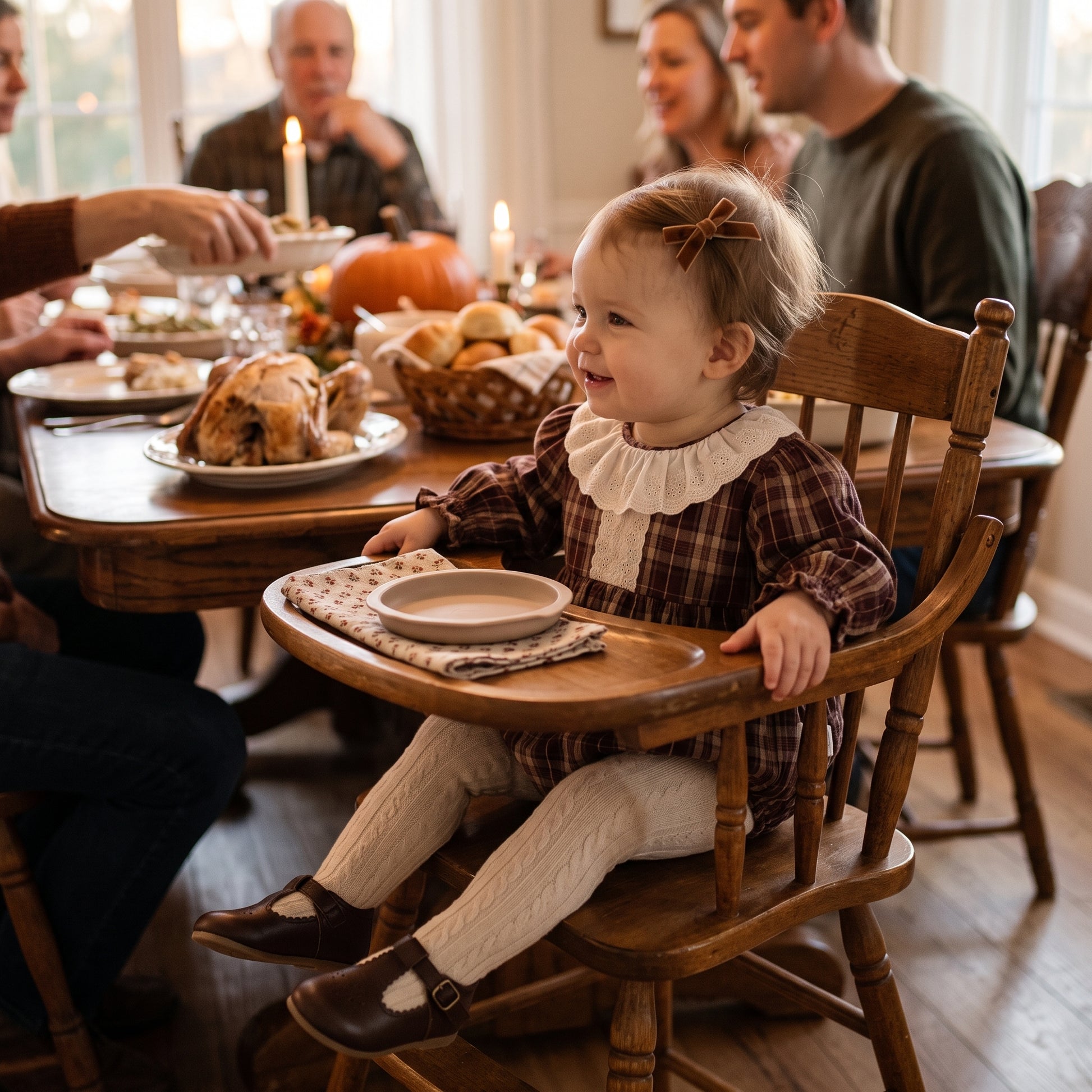 Baby girl sitting in a high chair wearing a brown plaid lace collar bubble romper at a family Thanksgiving dinner.