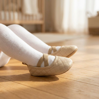 Side profile of an infant wearing Kidsely beige quilted ballerina shoes with white tights on a wooden floor.