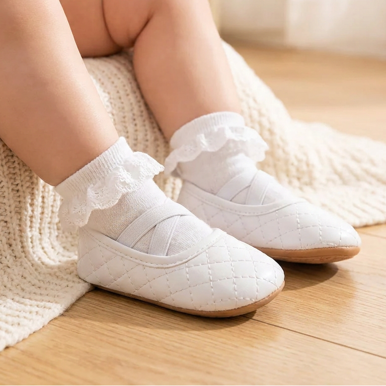 An infant sitting on a blanket wearing Kidsely white quilted ballerina shoes with ruffled white socks.