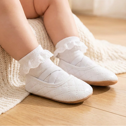 An infant sitting on a blanket wearing Kidsely white quilted ballerina shoes with ruffled white socks.